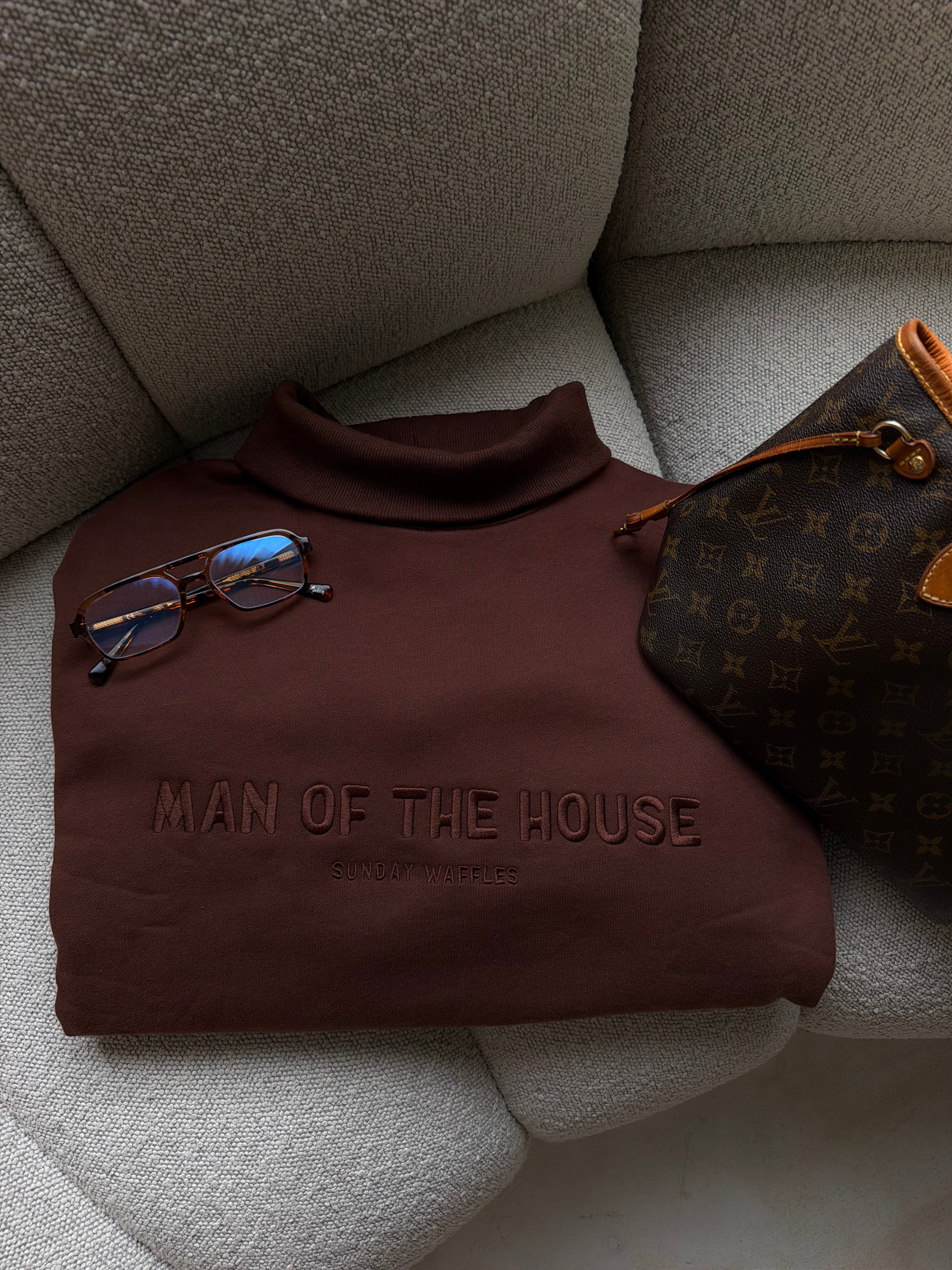 A folded brown turtleneck sweater with 'MAN OF THE HOUSE' embossed on the chest, styled on a gray textured sofa next to a Luxury bag and sunglasses.